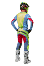 Piloto de motocross visto desde atrás con pantalones Techstar Ocuri 2024 y jersey a juego, azul claro, amarillo flúor y rojo, botas blancas y negras, casco negro con detalles neón, equipamiento coordinado de Alpinestars para carreras todoterreno