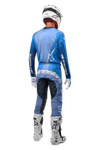 Piloto de motocross visto desde atrás con los pantalones Alpinestars Techstar Pneuma 2024 en azul marino y azul claro, combinados con un jersey azul a juego, casco blanco, guantes blancos y botas blancas, con la marca Alpinestars destacada en el asiento y las mangas.