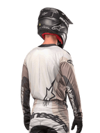 Piloto de motocross visto desde atrás con el Jersey Alpinestars Techstar Pneuma 2024 en gris arena y gris hierro, combinado con un casco negro con acabado de carbono, con un gran logo de estrella en la espalda y la marca Alpinestars en las mangas.