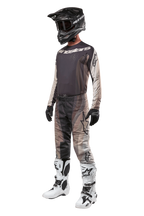 Pantalones Alpinestars Techstar Pneuma 2024, pantalones de motocross, color Dark Sand Iron Dust Gray, usados como parte de un conjunto completo de equipamiento con jersey a juego, casco negro, guantes negros y botas blancas, con patrones de camuflaje desértico en tonos similares y logotipos destacados