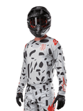 Alpinestars Techstar Rantera Jersey 2024, jersey de motocross en gris con patrones abstractos negros y acentos rojos, diseño de manga larga usado con casco y guantes negros, equipo técnico para carreras todoterreno