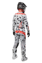 Piloto de motocross desde atrás con pantalones Alpinestars Techstar Rantera 2024, Haze Gray Camo con manchas negras y logotipos rojos, jersey a juego, casco negro y botas blancas, equipamiento completo de carreras todoterreno