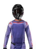 Alpinestars Techstar Ocuri Jersey 2024, jersey de motocross en púrpura y naranja intenso, usado por un piloto con casco y gafas negras, con perforaciones cortadas con láser en el pecho, ajuste atlético de carrera y logotipos rojos llamativos en los hombros y el pecho.