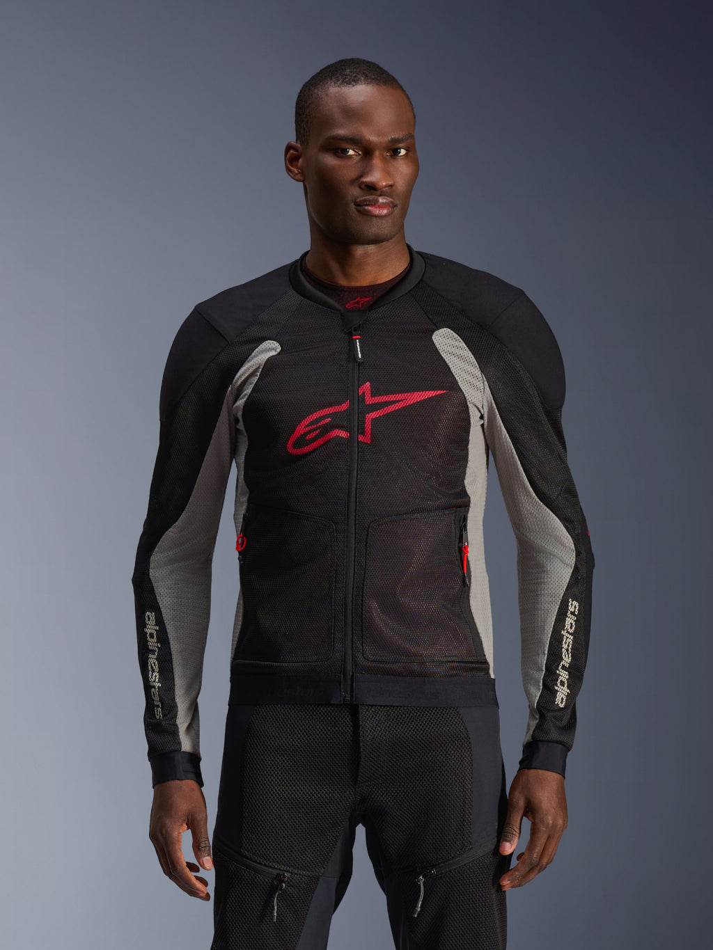 Chaqueta Alpinestars Troop-Air, chaqueta de malla para moto, negra y gris ceniza con detalles en rojo brillante, usada por un modelo, con paneles de malla técnica 2D transpirables para ventilación, un gran logotipo de Astars rojo en el pecho y un diseño técnico deportivo para conducción urbana.