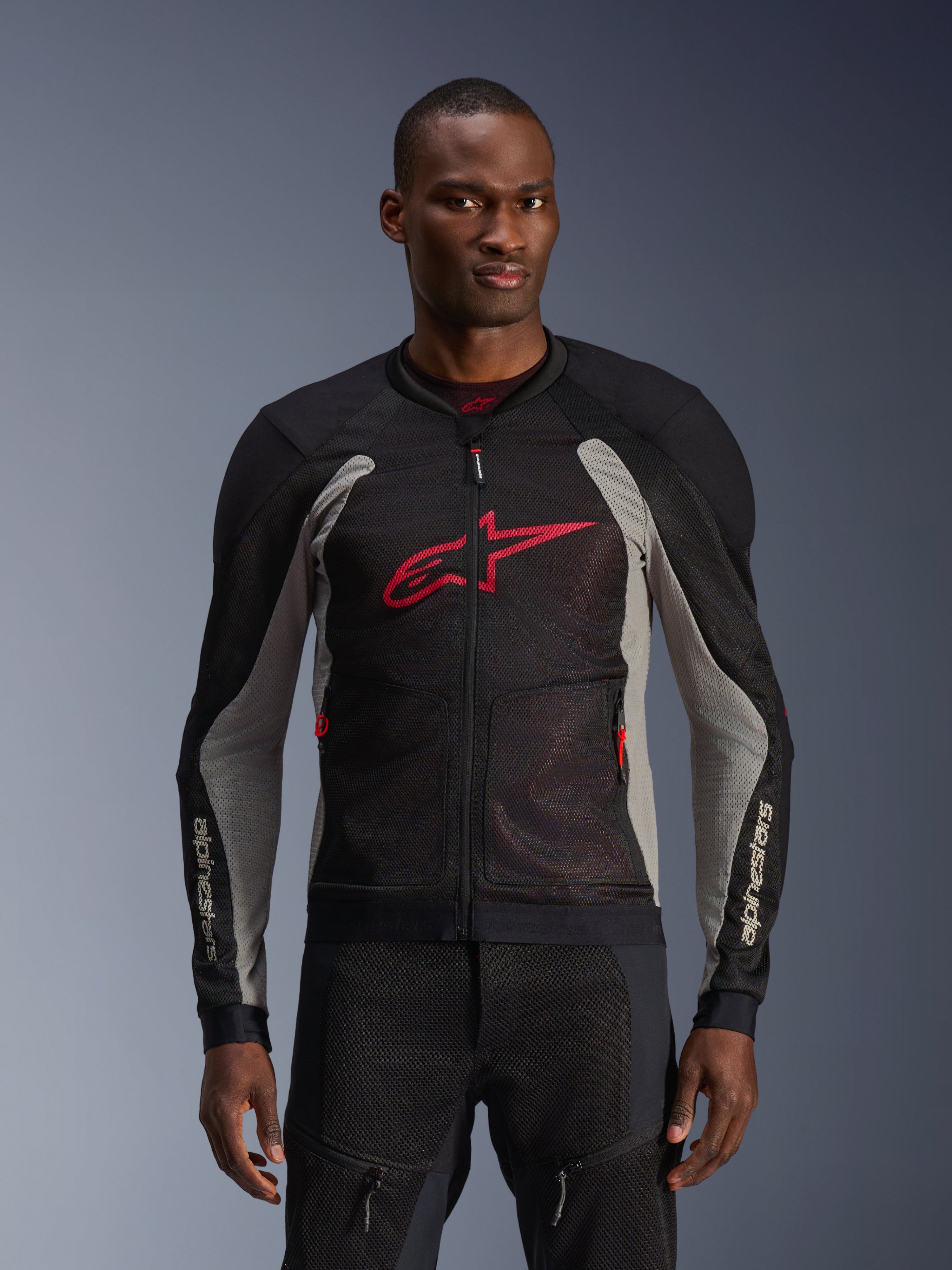 Chaqueta Alpinestars Troop-Air, chaqueta de malla para moto, negra y gris ceniza con detalles en rojo brillante, usada por un modelo, con paneles de malla técnica 2D transpirables para ventilación, un gran logotipo de Astars rojo en el pecho y un diseño técnico deportivo para conducción urbana.
