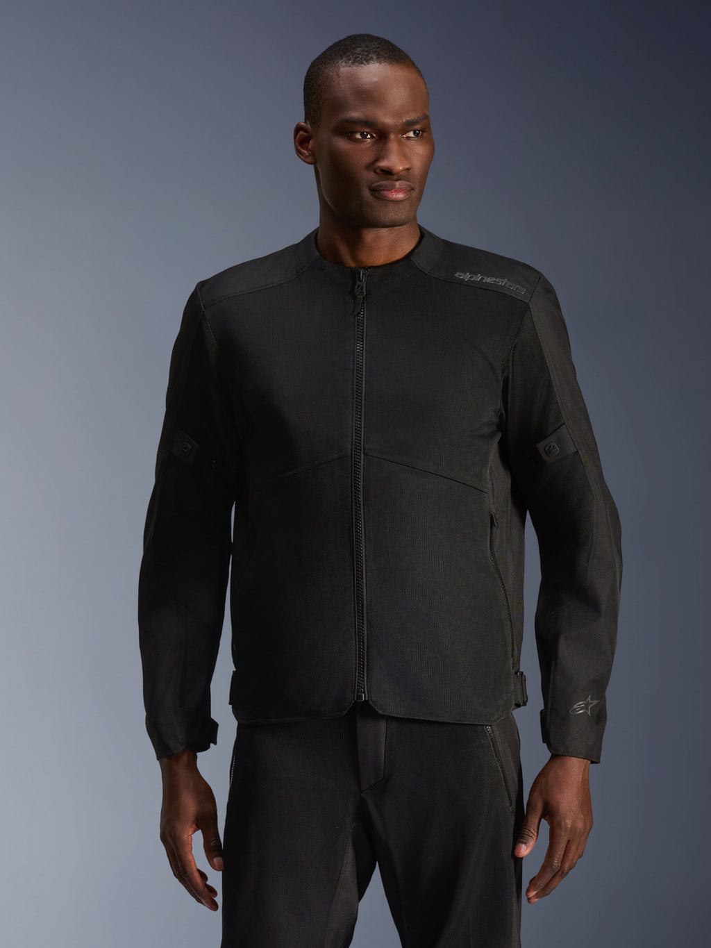 Modelo masculino con la chaqueta Alpinestars C-1 Air en negro, chaqueta de moto contemporánea con amplios paneles de malla técnica y cremallera frontal completa, combinada con pantalones de montar negros a juego, estética de estilo de vida urbano y desplazamientos
