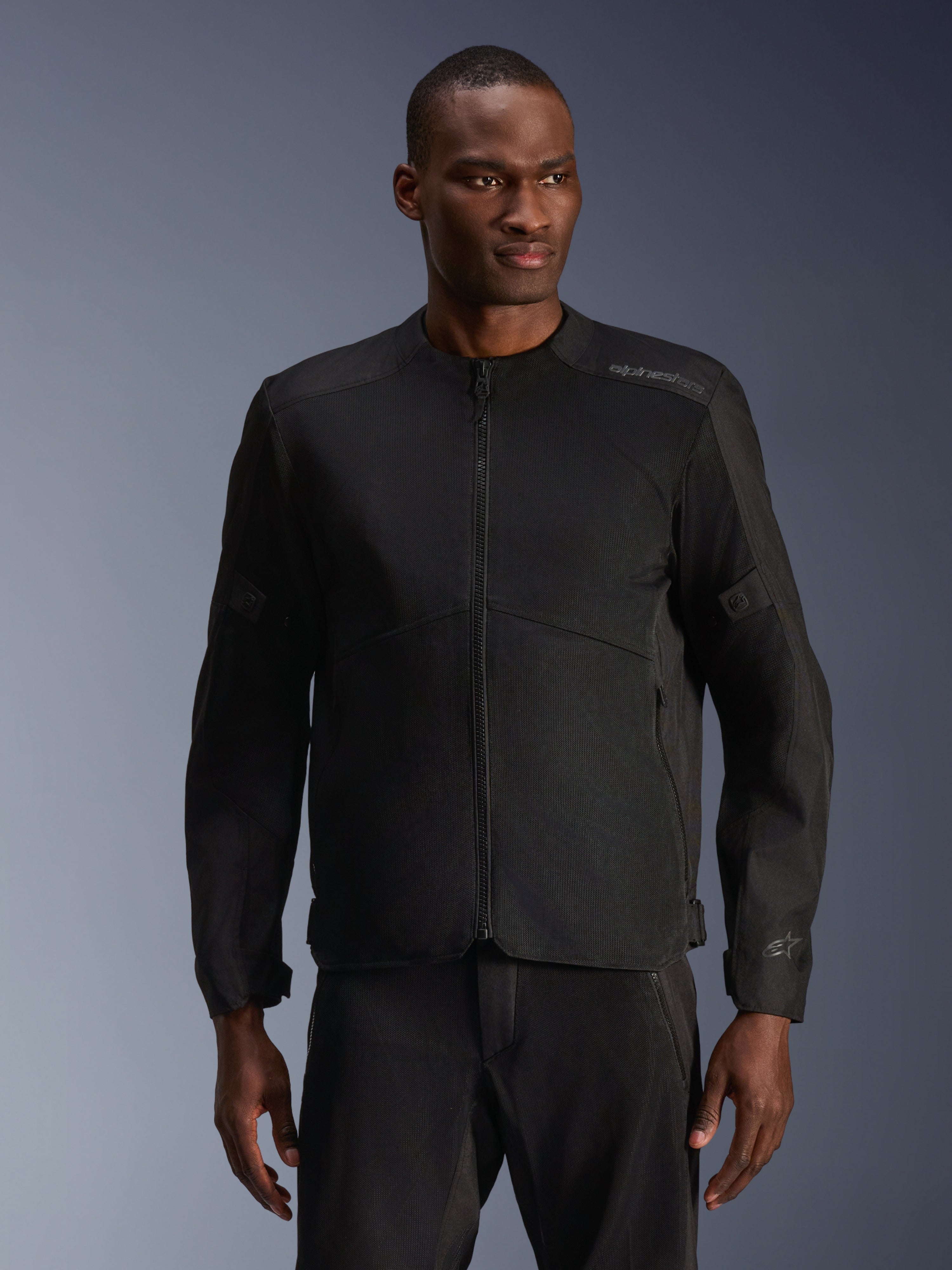 Modelo masculino con la chaqueta Alpinestars C-1 Air en negro, chaqueta de moto contemporánea con amplios paneles de malla técnica y cremallera frontal completa, combinada con pantalones de montar negros a juego, estética de estilo de vida urbano y desplazamientos