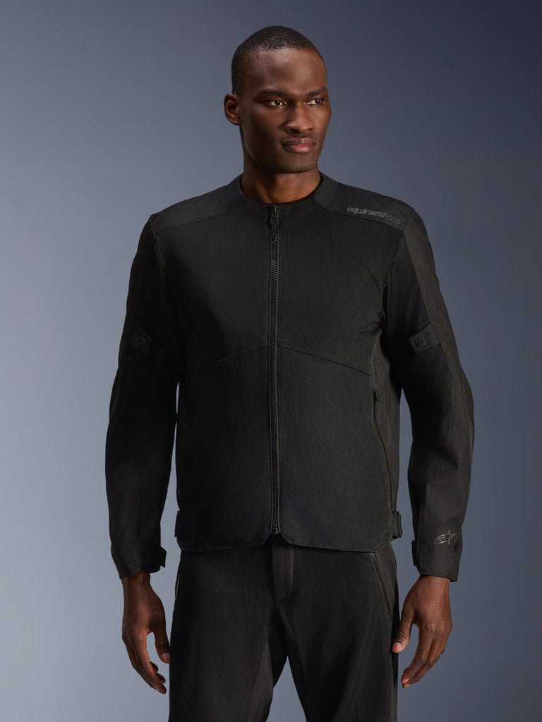Modelo masculino con la chaqueta Alpinestars C-1 Air en negro, chaqueta de moto contemporánea con amplios paneles de malla técnica y cremallera frontal completa, combinada con pantalones de montar negros a juego, estética de estilo de vida urbano y desplazamientos