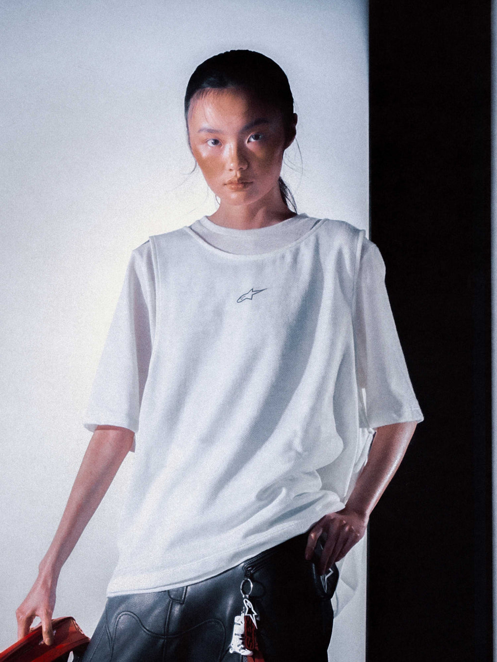 Alpinestars RSRV A1 SS Tee, camiseta blanca de manga corta con logo pequeño, estilo streetwear premium, persona usando pantalones negros de cuero con llavero, fondo degradado, iluminación dramática
