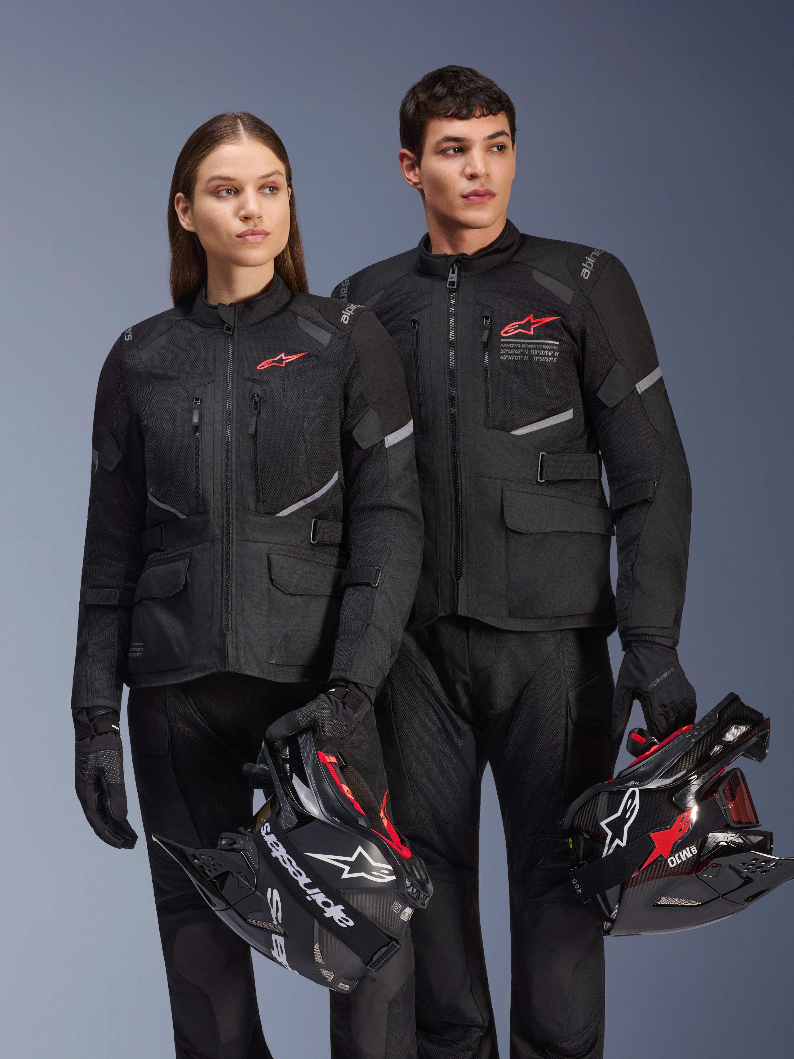 Hombre y mujer con chaquetas Alpinestars Andes Air Drystar®, equipamiento adventure touring negro con paneles de malla y detalles reflectantes, pantalones y guantes negros a juego, sosteniendo cascos de motocross, fotografía de estudio de ropa profesional para motociclistas
