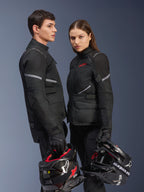 Pilotos de moto hombre y mujer con equipamiento Alpinestars, que incluye la chaqueta Stella Andes Air Drystar® en negro, pantalones textiles y guantes a juego, ambos sosteniendo cascos blancos y negros, toma de estudio que muestra ropa coordinada para adventure touring